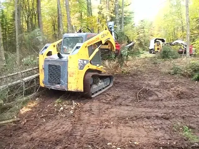 Two tracked skidsteers working a cleared, muddy lot lined with felled timber in autumn